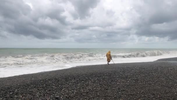 嵐の間に浜辺に金属探知機を持つ男 男はビーチで宝石を探しています 海岸の嵐警報中の嵐雲と大きな海の波 — ストック動画