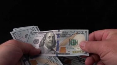 Adam siyah bir arka plan üzerinde dolar sayar. Bir adamın elinde bir demet Amerikan parası. Sahte bir banknot için 100 dolarlık banknotları kontrol ediyorum. Kavram: zenginlik, zenginleştirme, sahte para doğrulama.