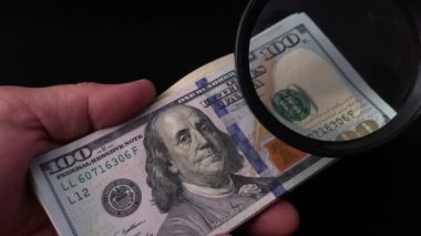 Bir adam büyüteçle dolarları kontrol eder. Mans elinde bir büyüteç ve yüz Amerikan doları. Sahte bir yüz dolar yüz dolarlık banknotları kontrol etmek.