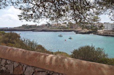 Taş bir duvar ve ağaçla çerçevelenmiş Akdeniz manzarası. Arka plandaki oteller.