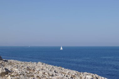 Okyanusta yelkenli, mavi gökyüzü, önünde kayalık kıyı şeridi.