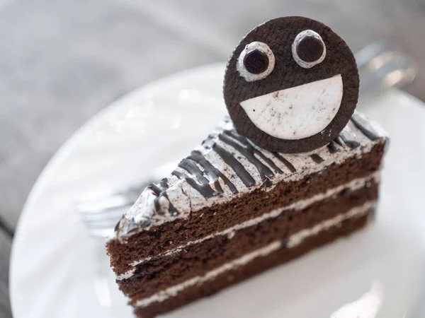 Piece of chocolate cake with face made from cookie and blurry pair of forks on white plate.