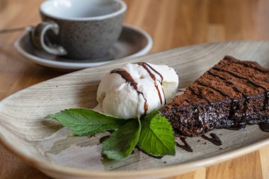 İştah açıcı koyu çikolatalı kekin dondurmalı dilimi ve arka planda odaklı kahve fincanı..