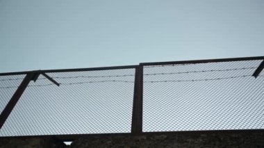Yüksek katlı beton çitler, tepesinde dikenli teller, sakin bir gökyüzüne karşı görülüyor..
