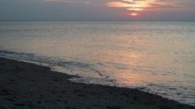 Kumsal ve gün batımında sakin bir deniz yüzeyi. Turuncu-pembe tonlarda deniz pelerini.