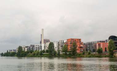 Nehrin kıyısındaki bir konut. Rahatla ve suyun üzerinde dinlen. Sakin nehir. Bulutlu gökyüzü. Suyun üzerindeki ağaçlar ve çalılar. Modern mimari. Renkli evler. Frankfurt Ben Main.