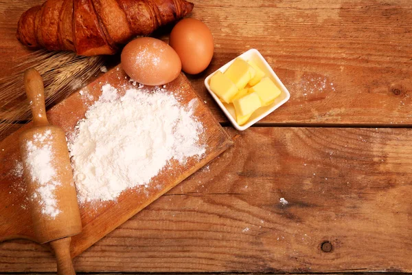 Wałek kuchenny, drewniana miska z mąką na drewnianym tle, jajka i masło do robienia rogalika na stole. — Zdjęcie stockowe
