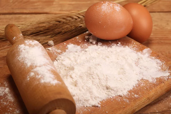 Kitchen rolling pin, wooden board with flour on wooden background, ears and eggs for making bread on table. — Stock Photo, Image