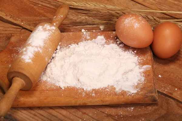 Kitchen rolling pin, wooden board with flour on wooden background, ears and eggs for making bread on table. — Stock Photo, Image