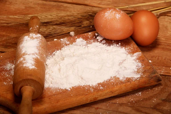 Kitchen rolling pin, wooden board with flour on wooden background, ears and eggs for making bread on table.