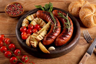 Ahşap masa üzerinde siyah tabakta sebze, baharat ve ekmek ile ızgara sosis. Makro görünümünü kapat.