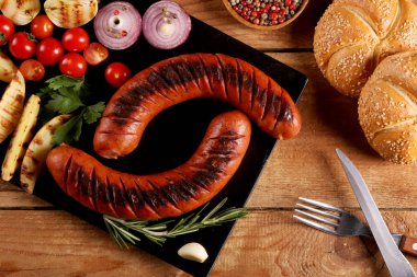 Ahşap masa üzerinde siyah tabakta sebze, baharat ve ekmek ile ızgara sosis. Makro görünümünü kapat.
