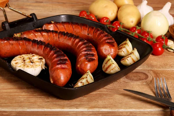 Grilled sausages with vegetables and spices in a pan on wooden background — Stock Photo, Image