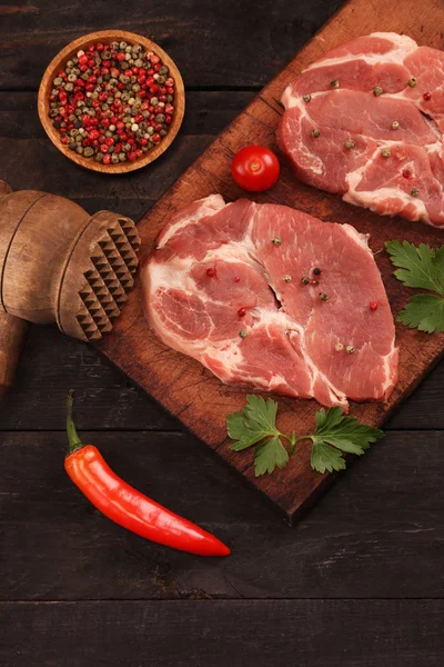 Carne cruda. Filete de cerdo crudo en una tabla de cortar con verduras, pimientos, tomate, sal y especias sobre un fondo negro — Foto de Stock