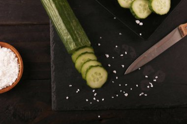 Siyah plaka ve siyah ahşap rustik masa üzerinde bıçak ve tuz ile dilimlenmiş yeşil salatalık. Üstte görüntü