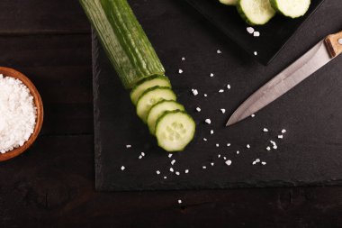 Siyah plaka ve siyah ahşap rustik masa üzerinde bıçak ve tuz ile dilimlenmiş yeşil salatalık. Üstte görüntü