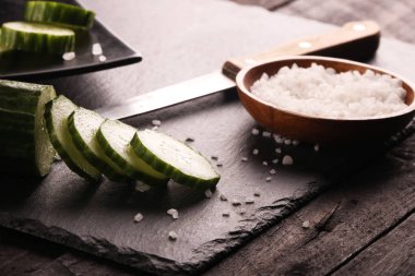 Siyah plaka ve siyah ahşap rustik masa üzerinde bıçak ve tuz ile dilimlenmiş yeşil salatalık.