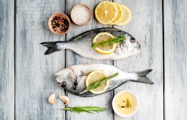 Ahşap arka planda biberiye dalları, limon dilimleri ve baharatlar ile servis edilen ham deniz bas 