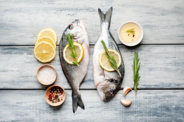 Ahşap arka planda biberiye dalları, limon dilimleri ve baharatlar ile servis edilen ham deniz bas 