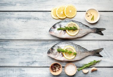 Ahşap arka planda biberiye dalları, limon dilimleri ve baharatlar ile servis edilen ham deniz bas 