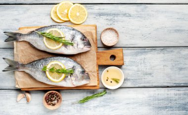 Ahşap arka planda biberiye dalları, limon dilimleri ve baharatlar ile servis edilen ham deniz bas 
