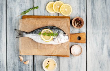 Ahşap arka planda biberiye dalları, limon dilimleri ve baharatlar ile servis edilen ham deniz bas 