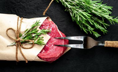 Çiğ sığır eti biftek taş arka plan üzerinde biberiye dalları ile zanaat kağıt sarılmış 