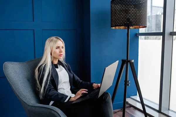 Elegant businesswoman sitting in armchair and working on laptop in blue interior