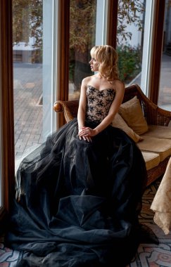 Pencerenin yanında poz veren zarif gece elbisesi giymiş genç sarışın kadın.