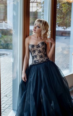 Pencerenin yanında poz veren zarif gece elbisesi giymiş genç sarışın kadın.