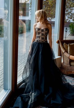 Pencerenin yanında poz veren zarif gece elbisesi giymiş genç sarışın kadın.