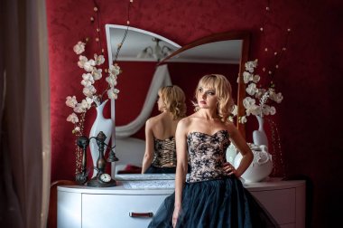 Pencerenin yanında poz veren zarif gece elbisesi giymiş genç sarışın kadın.