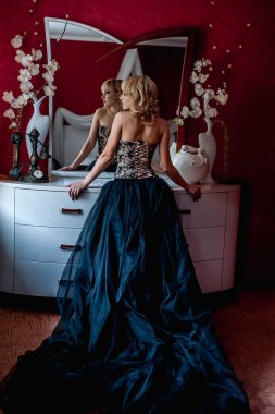 Zarif gece elbisesi giymiş genç sarışın kadın aynanın önünde poz veriyor. 