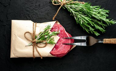 Çiğ sığır eti biftek taş arka plan üzerinde biberiye dalları ile zanaat kağıt sarılmış 