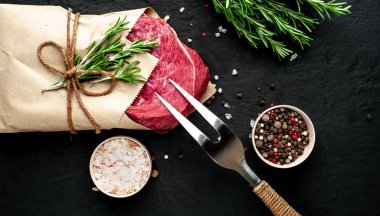 Çiğ sığır eti biftek taş arka plan üzerinde biberiye dalları ve baharatlar ile zanaat kağıt sarılmış 