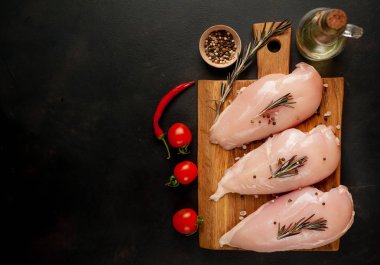 Ahşap doğrama tahtasında tuz, biberiye dalları ve zeytinyağı şişesi ile çiğ tavuk fileto