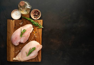 Ahşap doğrama tahtasında tuz, biberiye dalları ve zeytinyağı şişesi ile çiğ tavuk fileto