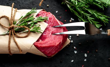 Çiğ sığır eti biftek taş arka plan üzerinde biberiye dalları ve baharatlar ile zanaat kağıt sarılmış 