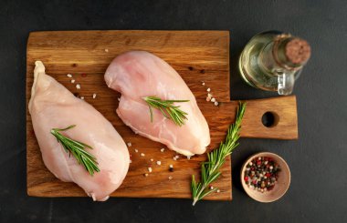 Ahşap doğrama tahtasında tuz, biberiye dalları ve zeytinyağı şişesi ile çiğ tavuk fileto
