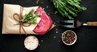 Çiğ sığır eti biftek taş arka plan üzerinde biberiye dalları ve baharatlar ile zanaat kağıt sarılmış 