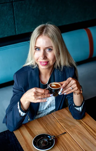 Stylish blonde woman drinking cup of coffee in blue cafe interior