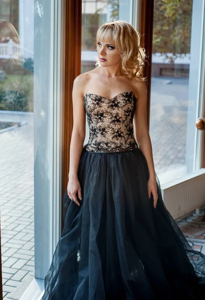 Pencerenin yanında poz veren zarif gece elbisesi giymiş genç sarışın kadın.