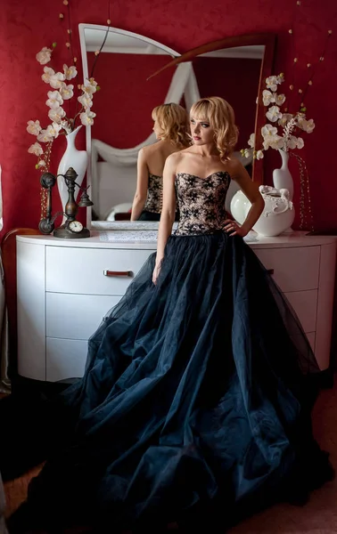 Pencerenin yanında poz veren zarif gece elbisesi giymiş genç sarışın kadın.