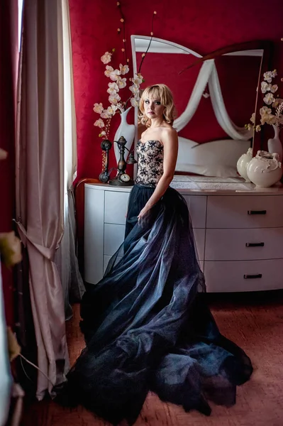 Pencerenin yanında poz veren zarif gece elbisesi giymiş genç sarışın kadın.