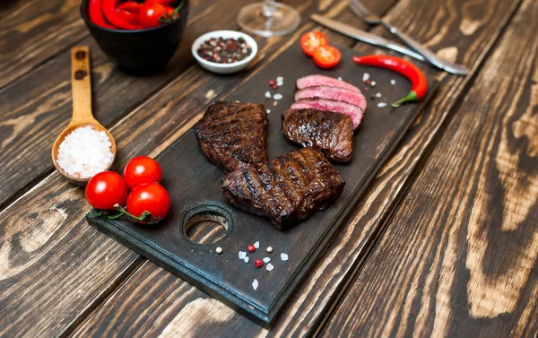 茶色の木製のテーブルの上に木製のボード上にチェリートマトと唐辛子と新鮮なグリルビーフステーキ — ストック写真