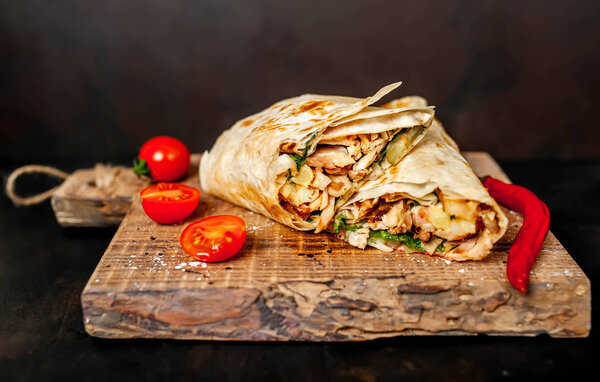 Traditional mexican burrito wrapped with chicken and vegetables served with ripe tomatoes on wooden chopping board 