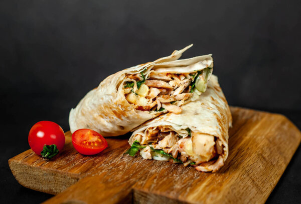Traditional mexican burrito wrapped with chicken and vegetables served with ripe tomatoes on wooden chopping board 