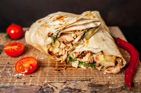 Traditional mexican burrito wrapped with chicken and vegetables served with ripe tomatoes on wooden chopping board 