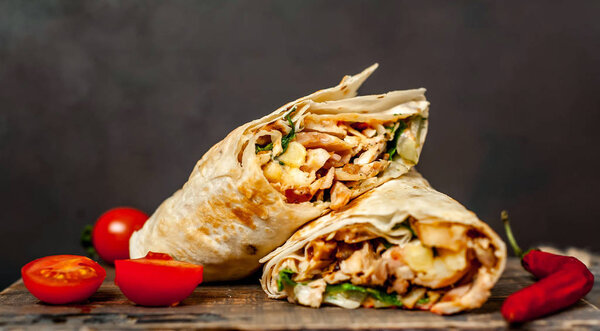 Traditional mexican burrito wrapped with chicken and vegetables served with ripe tomatoes on wooden chopping board 