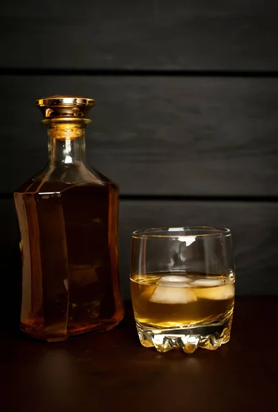 Glass with icy whiskey and bottle on dark wooden background